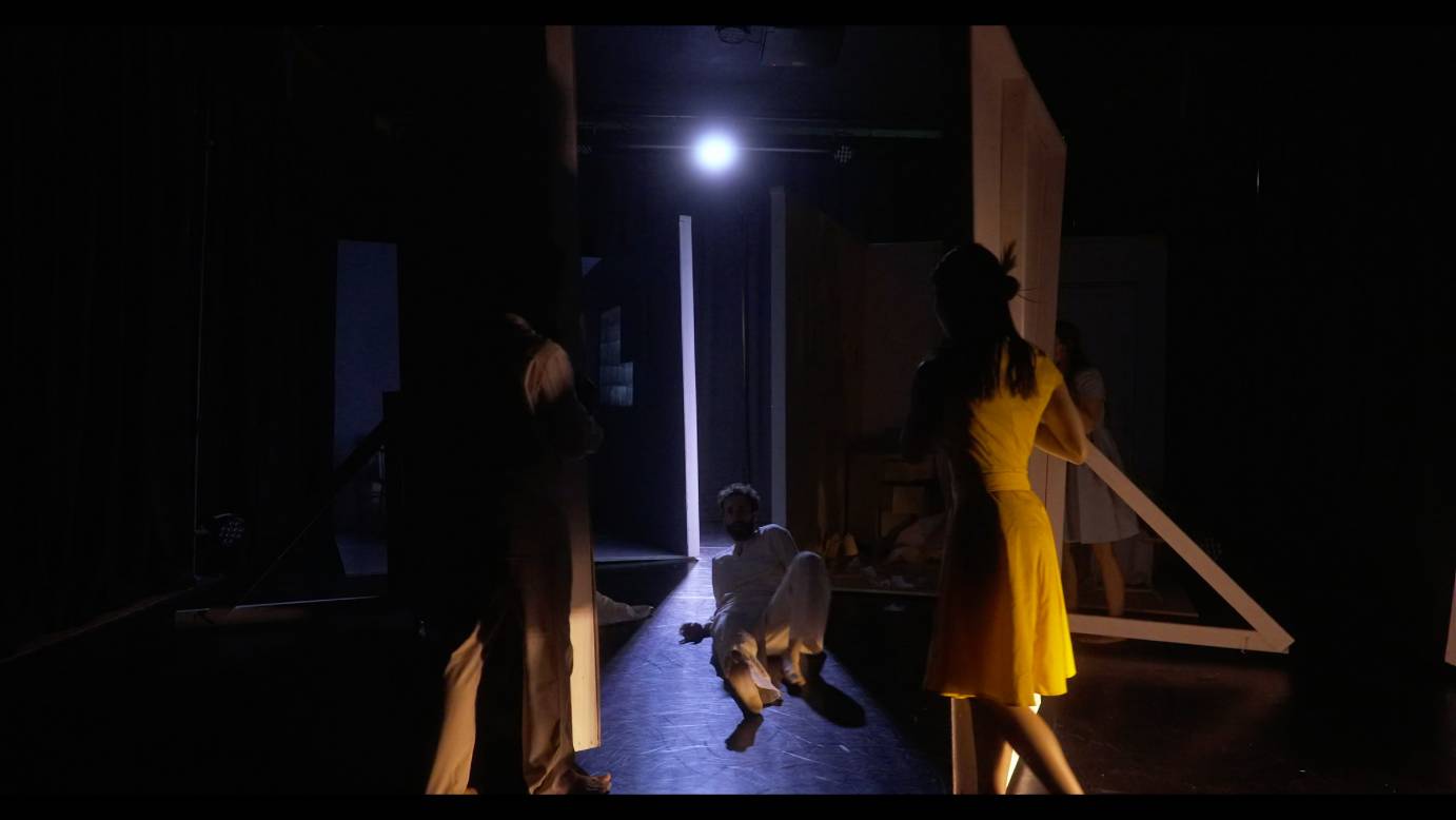 lit by a sight light a male dancer lies in a stripe of light on the floor as he is stared at by a man and woman to each side of him.